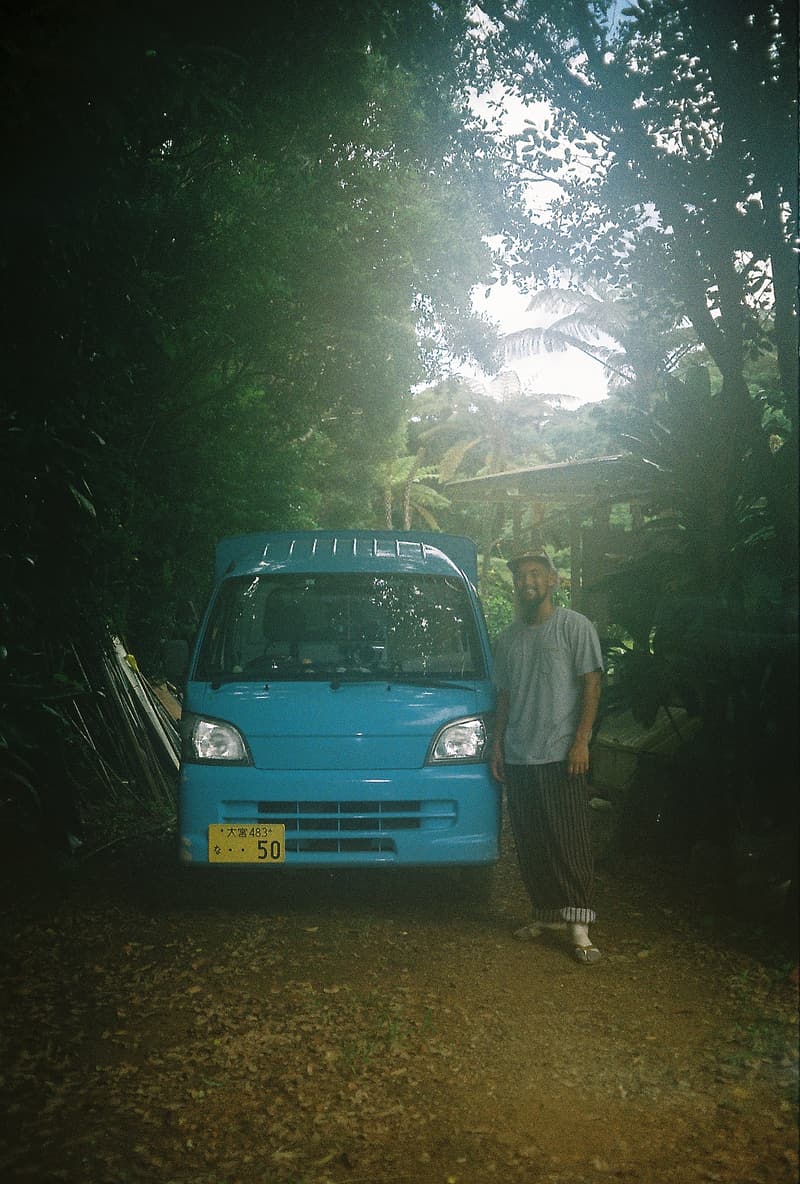 A man beside a small blue kei truck in Okinawan greenery